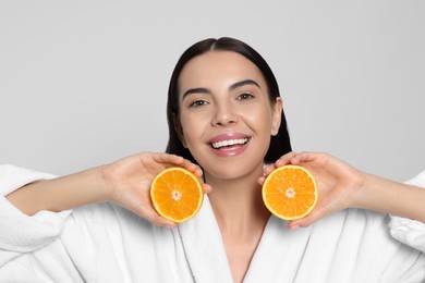 Woman in bathrobe holding juicy cut orange on light grey background. Spa treatment
