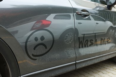 Phrase Wash Me and sad emoticon drawn on dirty car, closeup