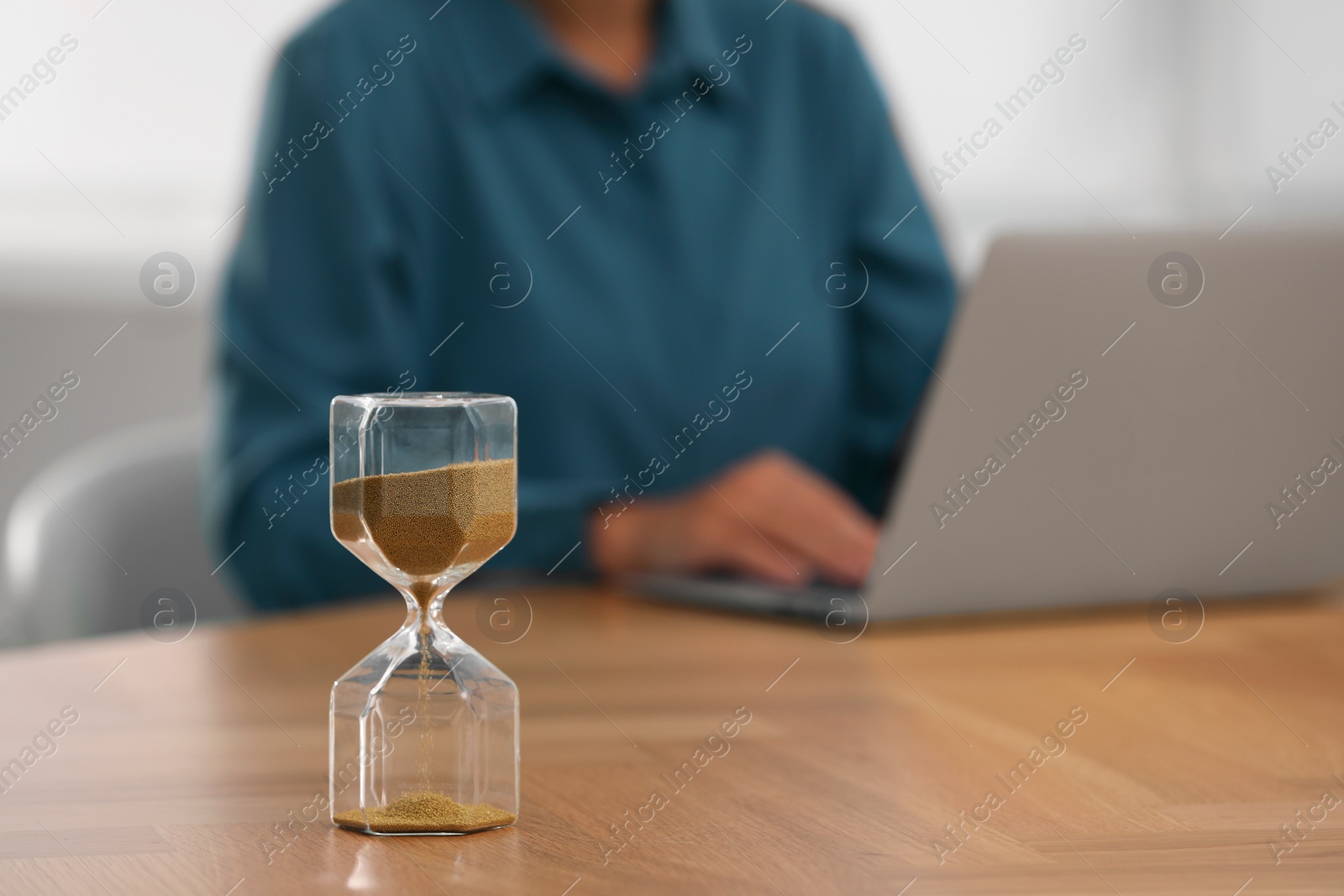 Photo of Hourglass with flowing sand on table. Woman using laptop indoors, selective focus