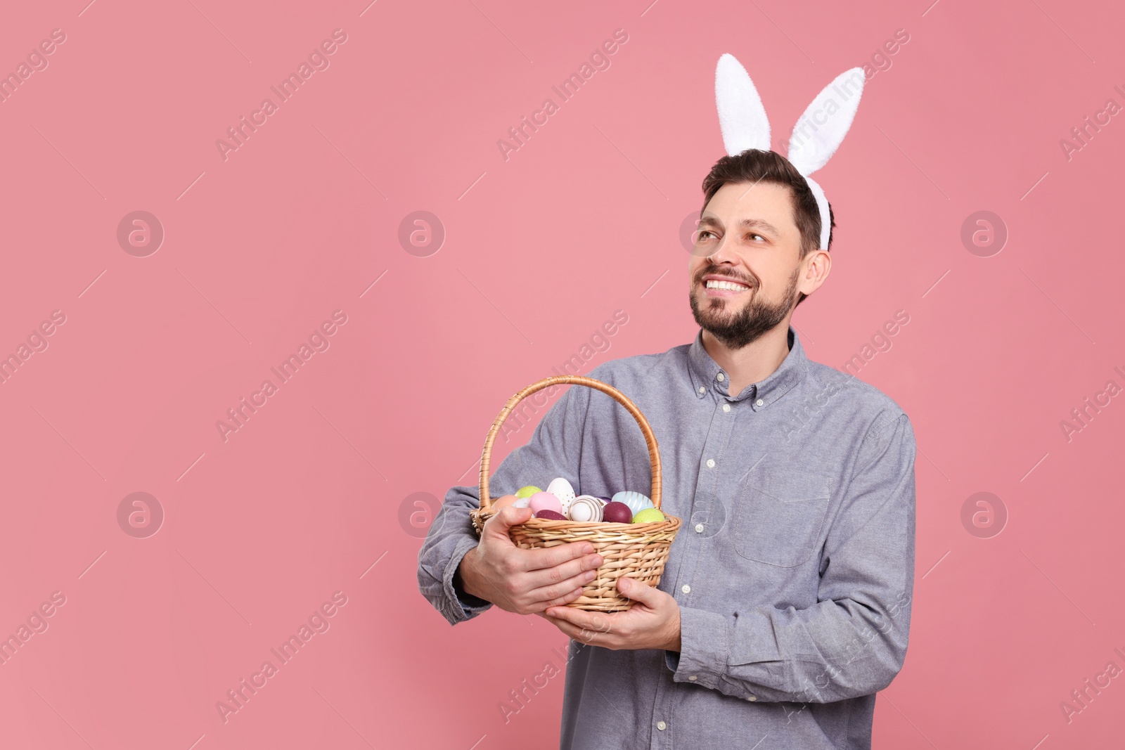 Photo of Happy man in bunny ears headband holding wicker basket with painted Easter eggs on pink background. Space for text