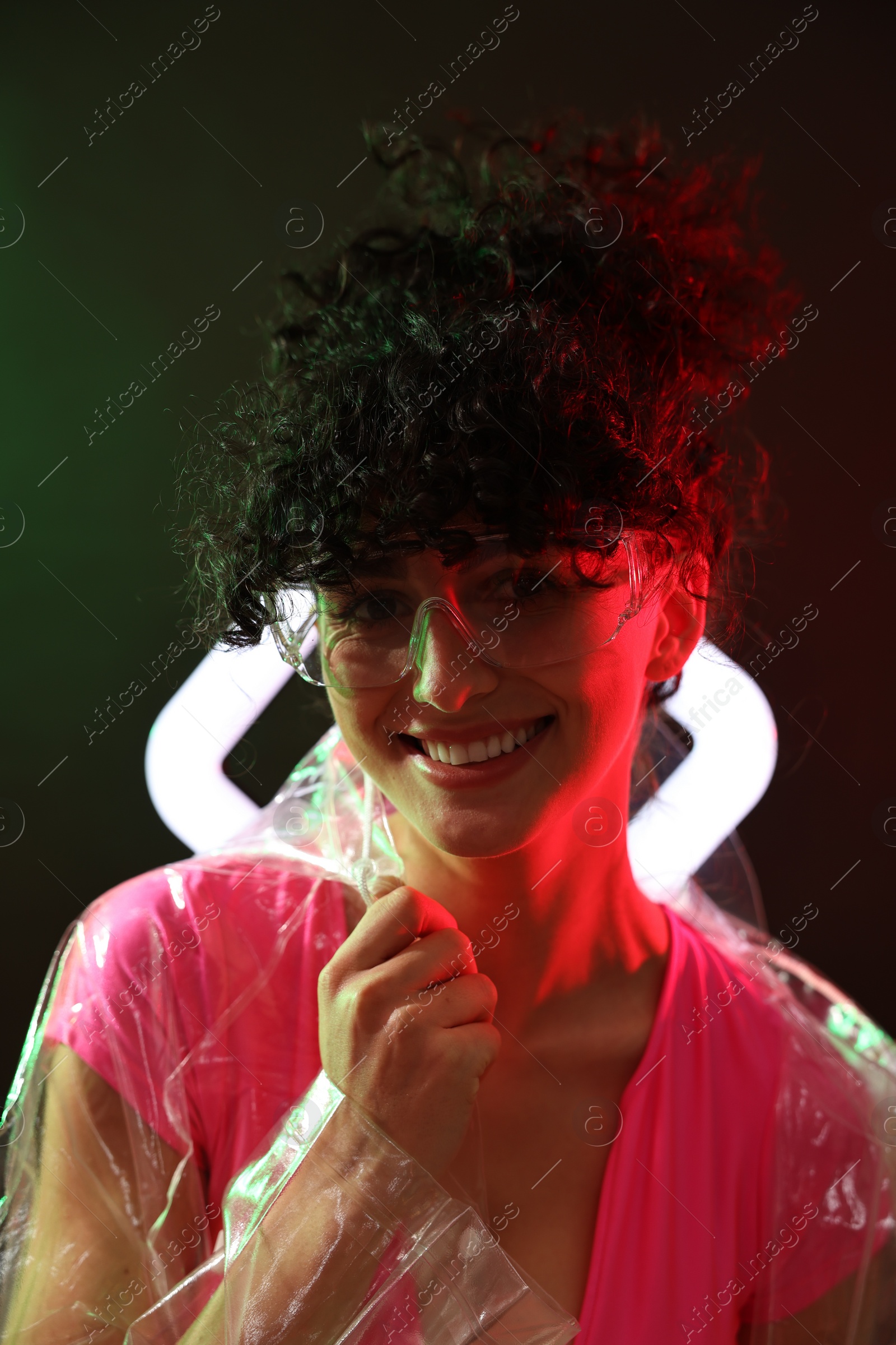 Photo of Beautiful young woman in transparent coat and sunglasses with square lamp on color background in neon lights