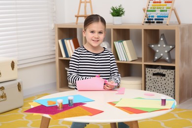 Cute little girl cutting pink paper at desk in room. Home workplace