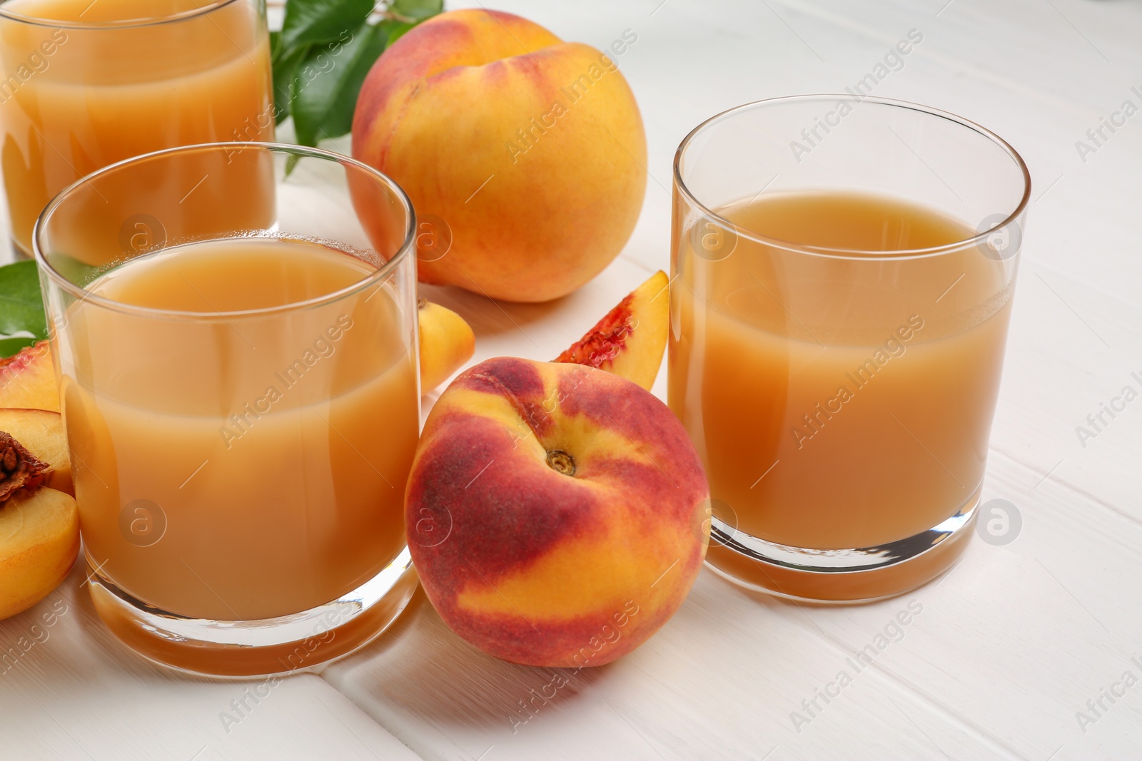 Photo of Glasses of peach juice, fresh fruits and leaves on white wooden table, closeup
