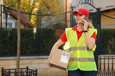 Photo of Courier in uniform with parcel talking on smartphone near private house outdoors, space for text