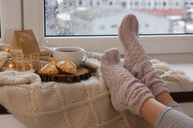 Photo of Woman in knitted socks relaxing near window at home, closeup
