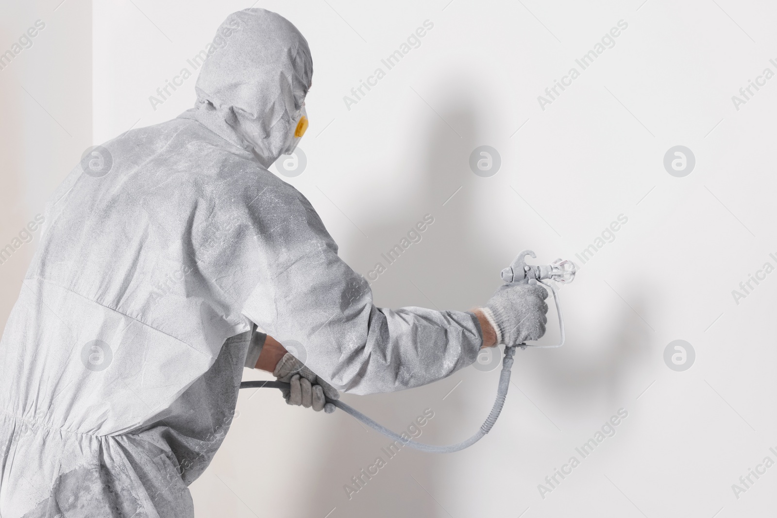 Photo of Decorator in protective overalls painting wall with spray gun indoors