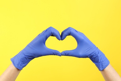 Person in medical gloves showing heart gesture on yellow background, closeup of hands