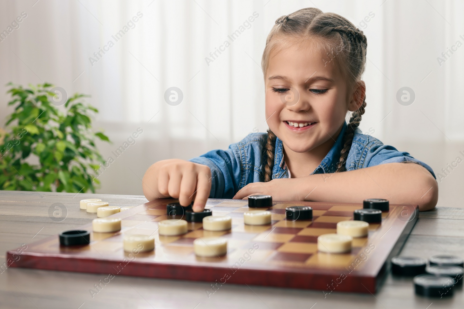 Photo of Playing checkers. Little girl thinking about next move at table in room