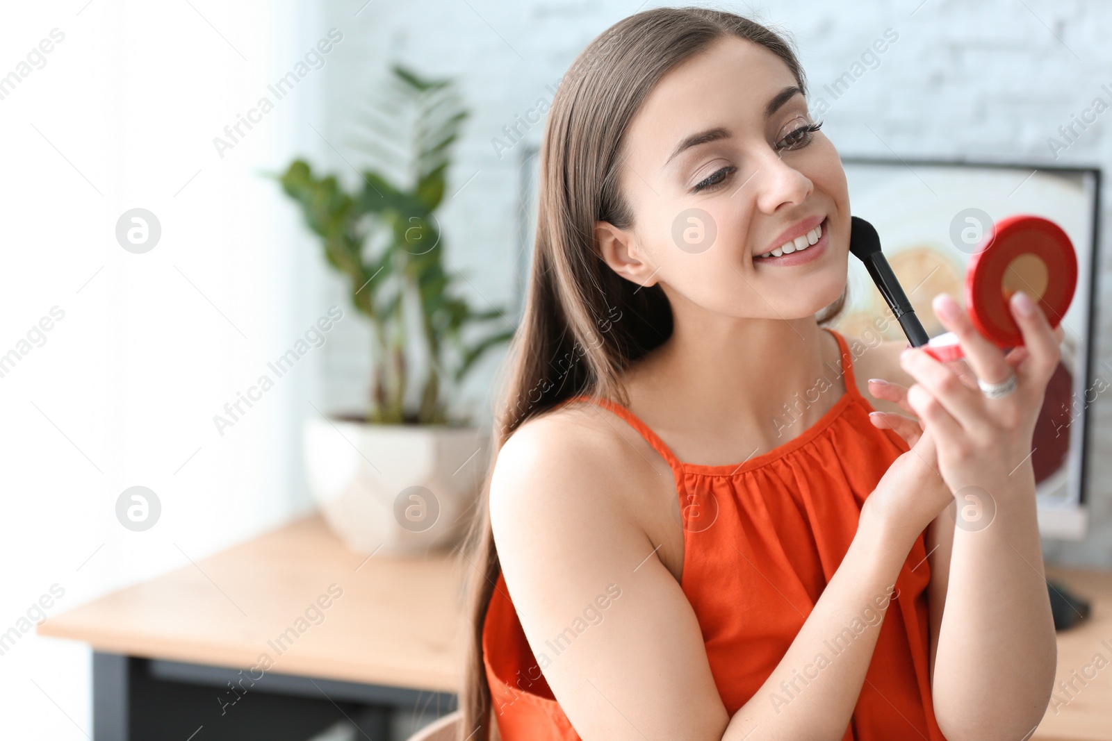 Photo of Young beautiful woman holding mirror while applying makeup in room
