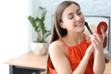 Young beautiful woman holding mirror while applying makeup in room