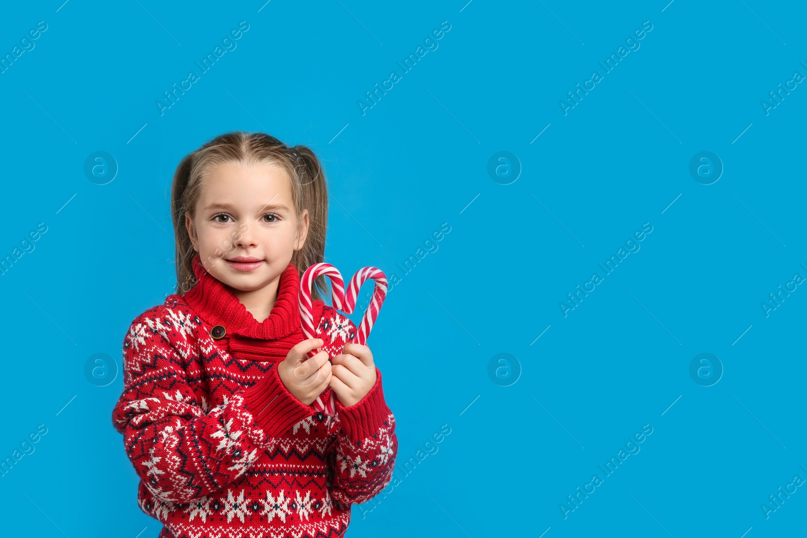 Photo of Cute little girl in knitted Christmas sweater holding candy canes on blue background, space for text