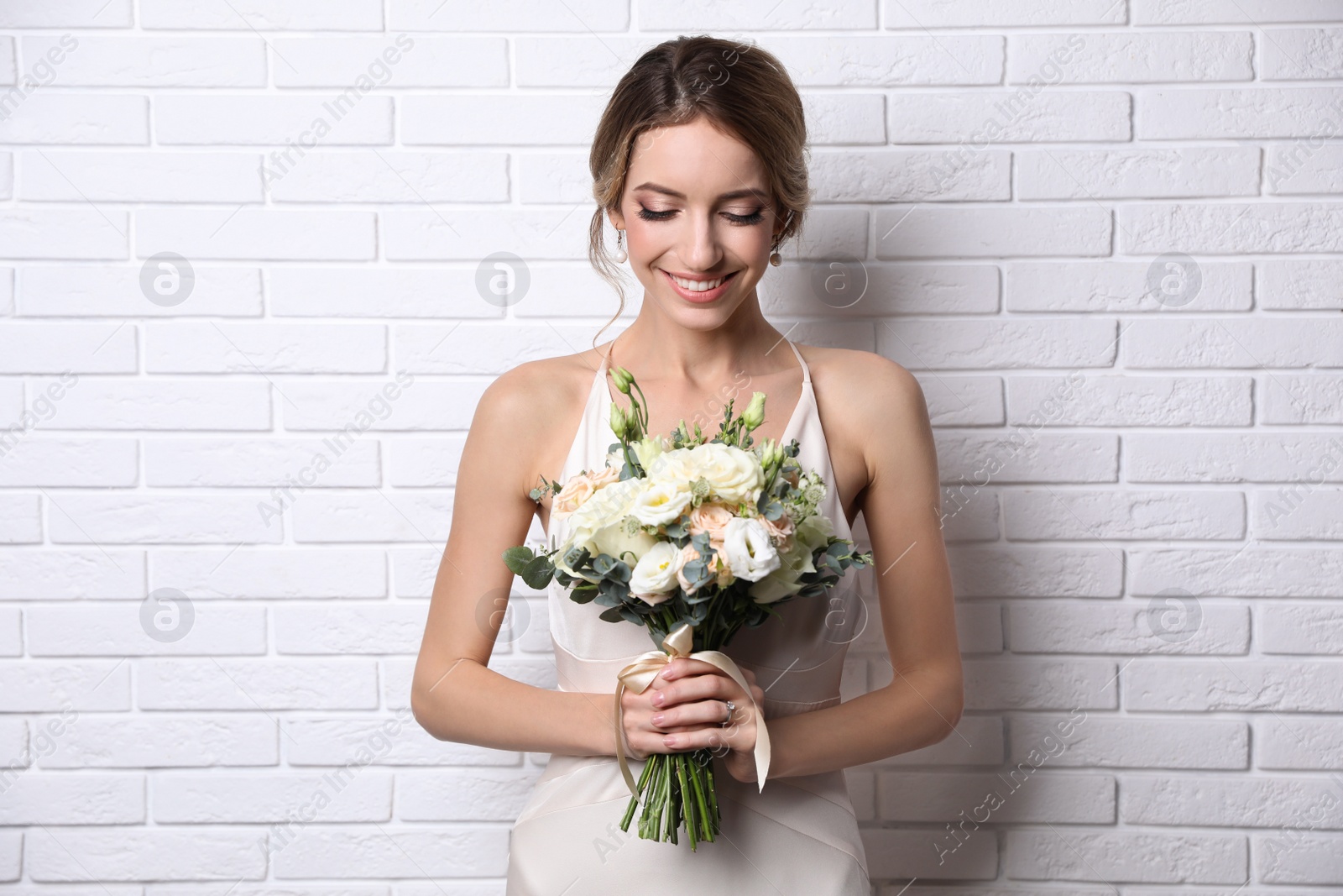 Photo of Young bride with elegant hairstyle holding wedding bouquet near white brick wall