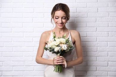 Young bride with elegant hairstyle holding wedding bouquet near white brick wall