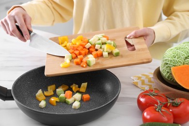 Photo of Woman pouring mix of cut vegetables into frying pan at table, closeup