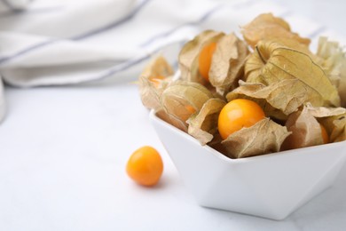 Ripe physalis fruits with calyxes in bowl on white marble table, closeup. Space for text