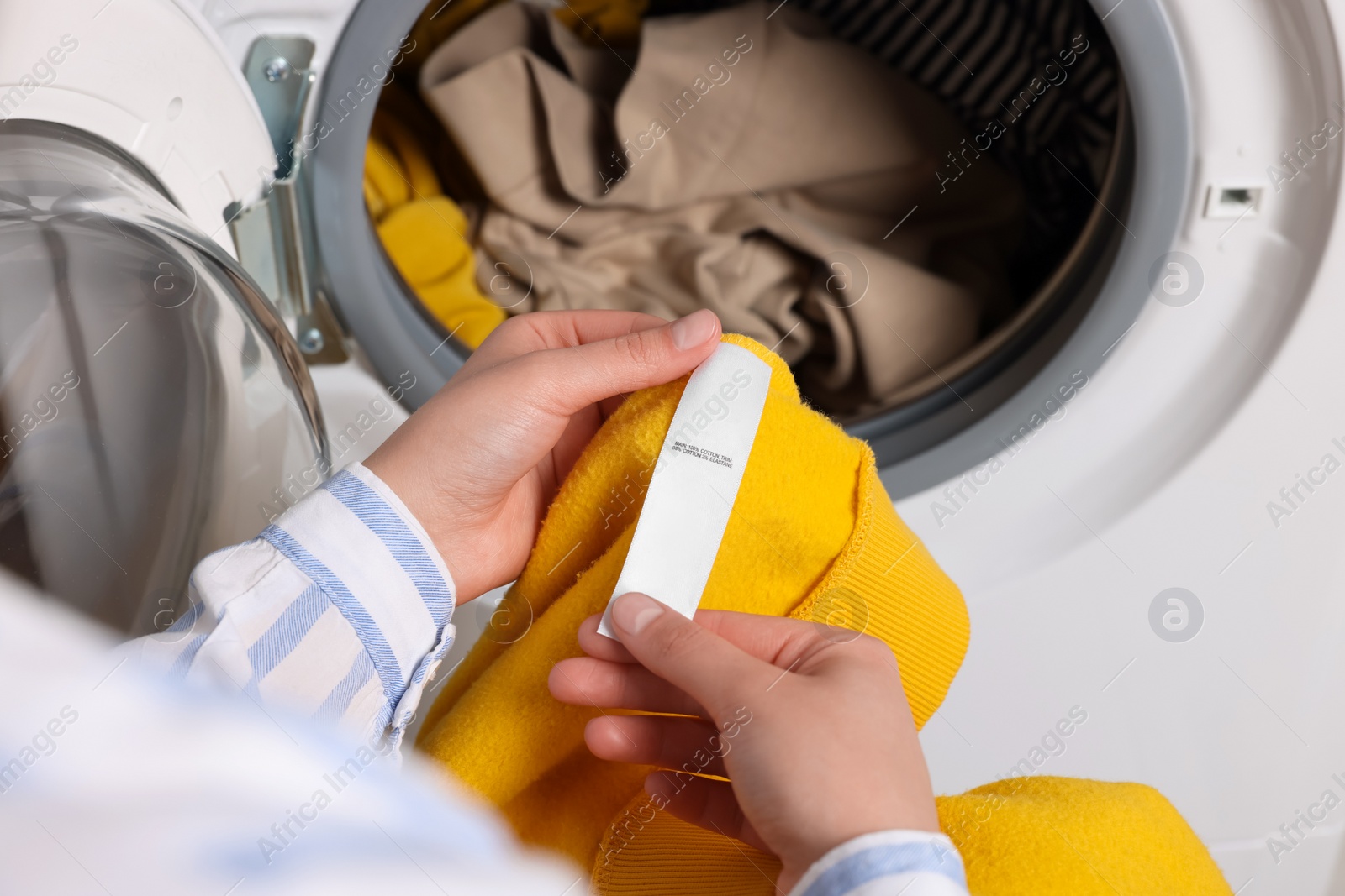 Photo of Woman reading clothing label with care symbols and material content on yellow shirt near washing machine, closeup