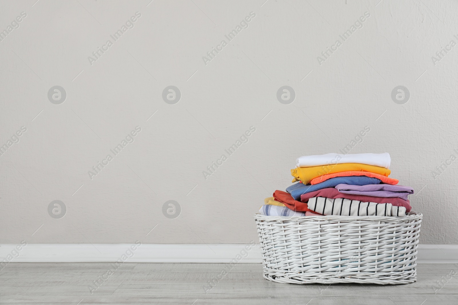 Photo of Wicker laundry basket with clean clothes on floor near light wall. Space for text