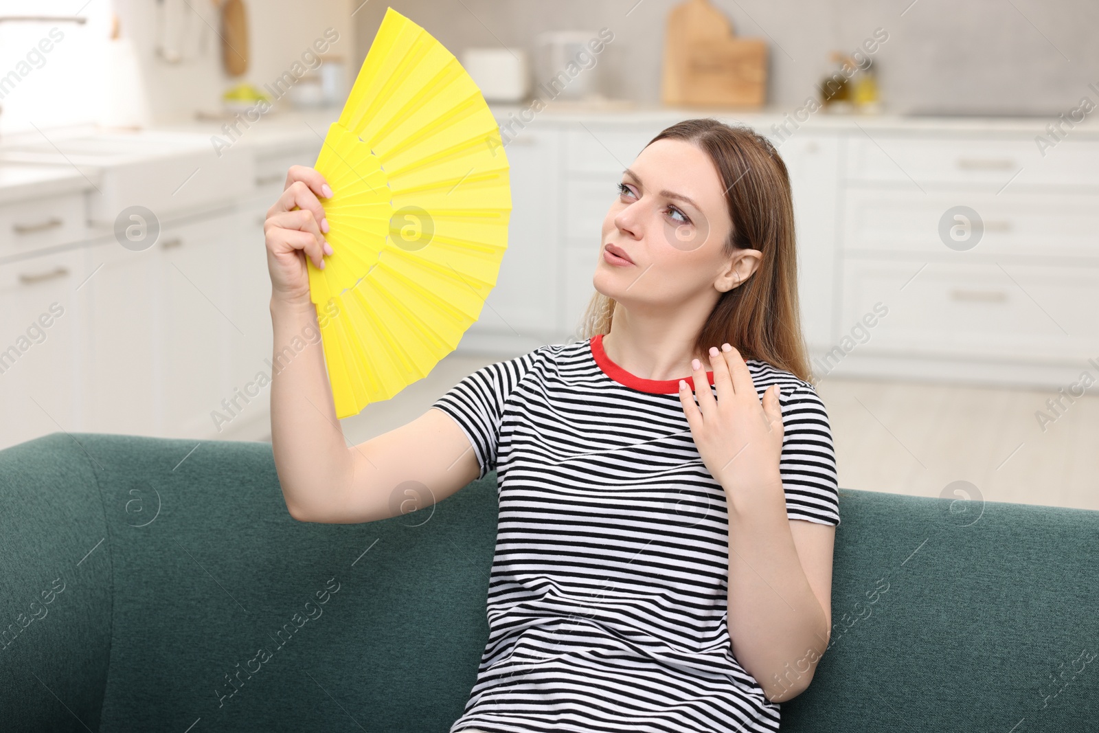 Photo of Woman waving yellow hand fan to cool herself on sofa at home