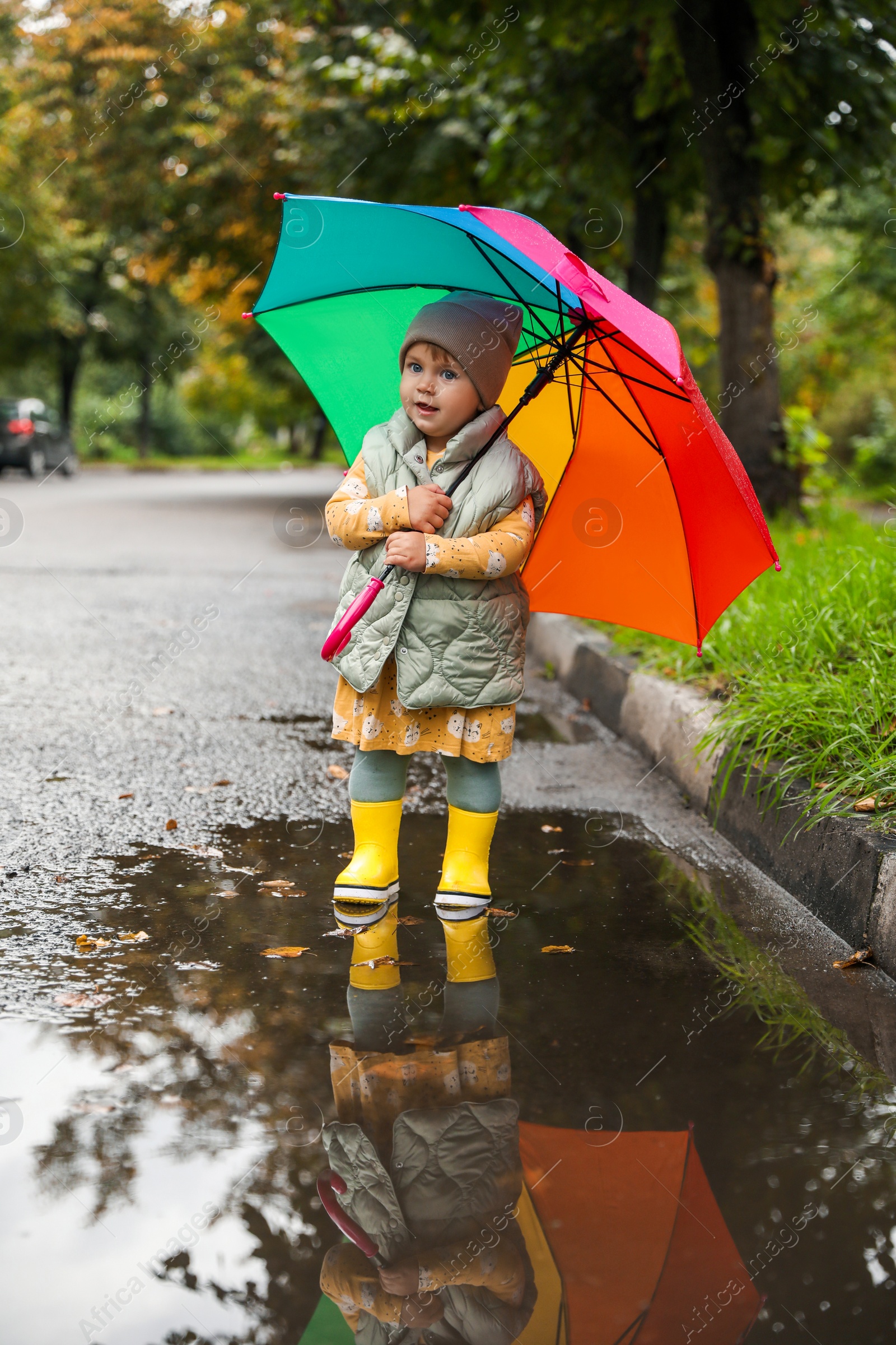 Photo of Cute little girl with colorful umbrella standing in puddle outdoors