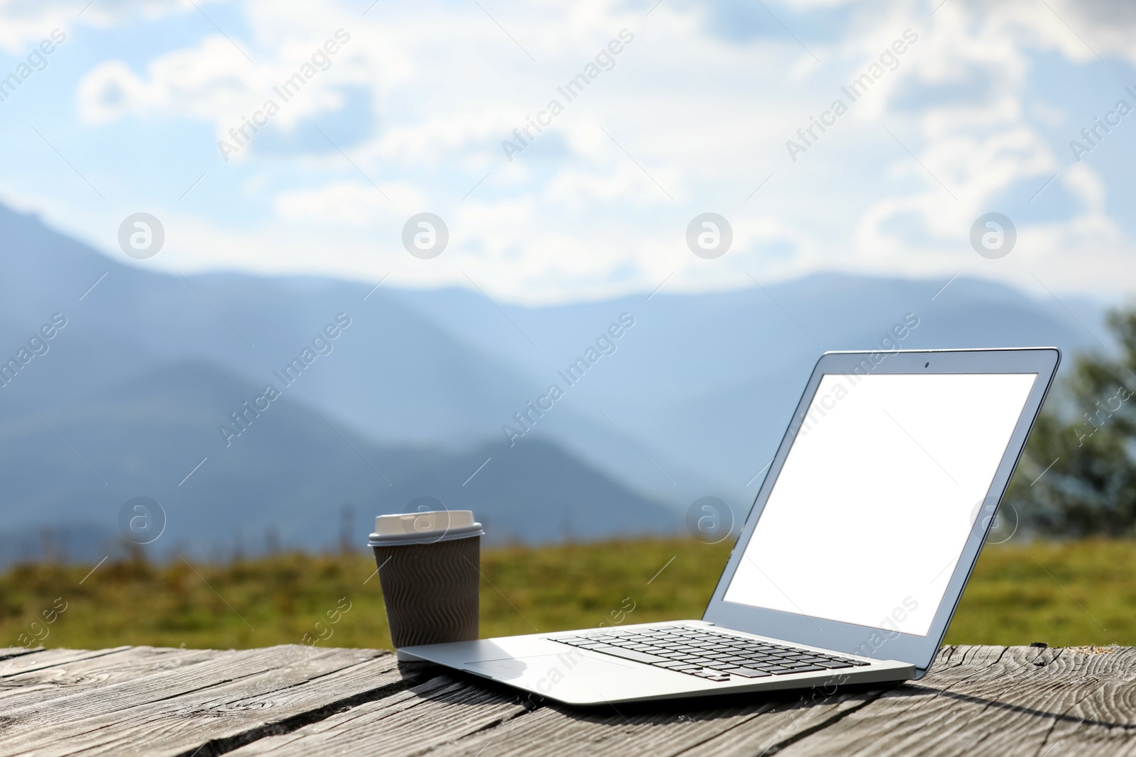 Photo of Modern laptop with blank screen and coffee cup on wooden surface in mountains, space for text. Working outdoors