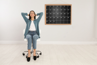 Beautiful young woman sitting near board calendar indoors