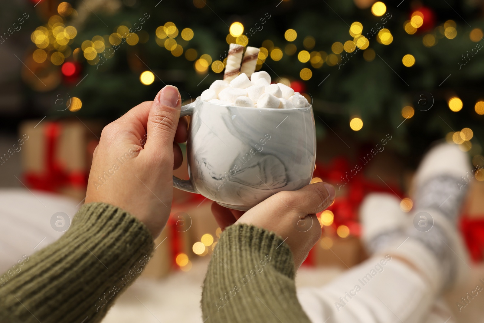 Photo of Woman holding cup of delicious cocoa with marshmallows near Christmas tree at home, closeup. Space for text