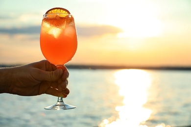 Man with glass of fresh summer cocktail near water at sunset, closeup. Space for text