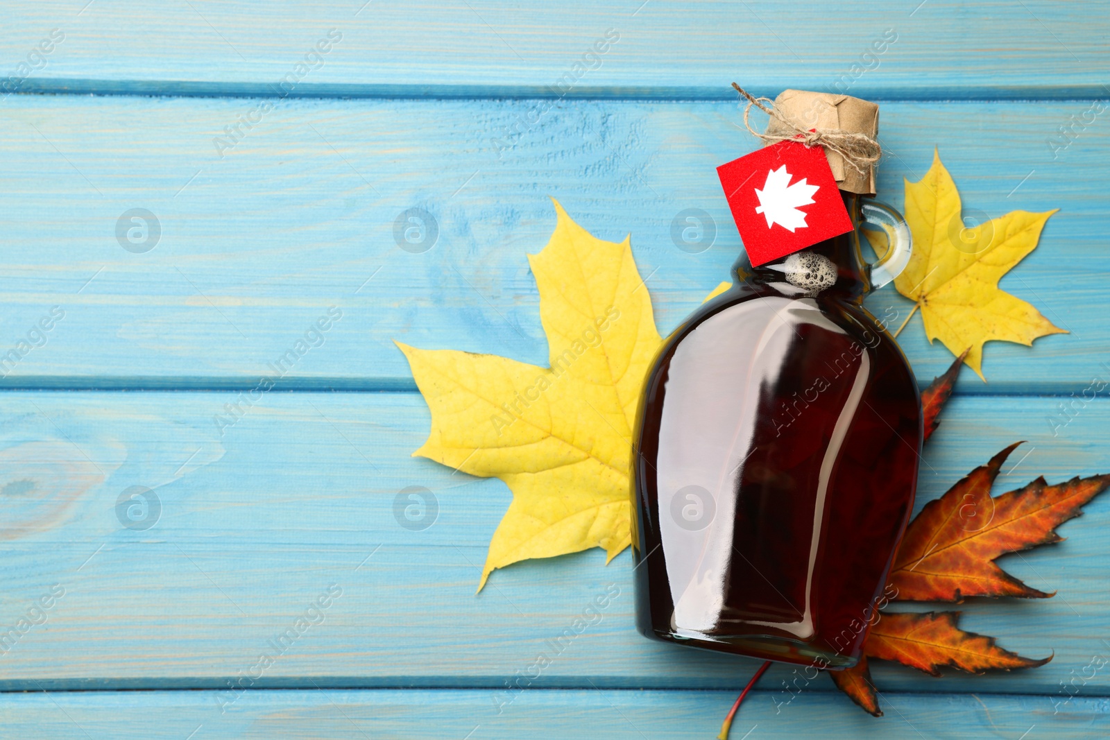 Photo of Glass bottle of tasty maple syrup and dry leaves on light blue wooden table, flat lay. Space for text