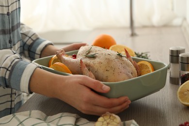 Photo of Woman holding baking pan with Chicken and orange slices at wooden table, closeup