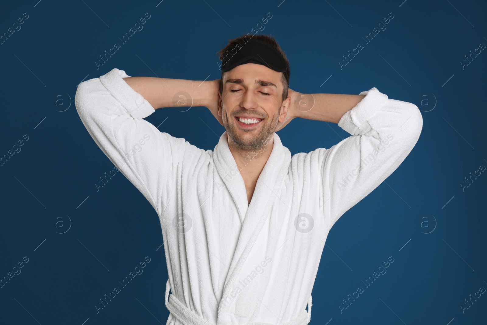 Photo of Happy young man in bathrobe and eye sleeping mask stretching on blue background