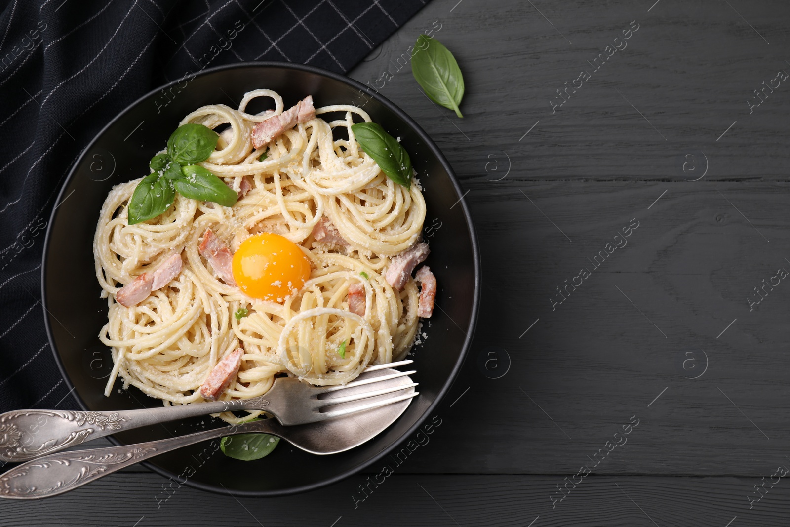 Photo of Bowl of delicious pasta Carbonara with egg yolk and cutlery on black wooden table, flat lay. Space for text