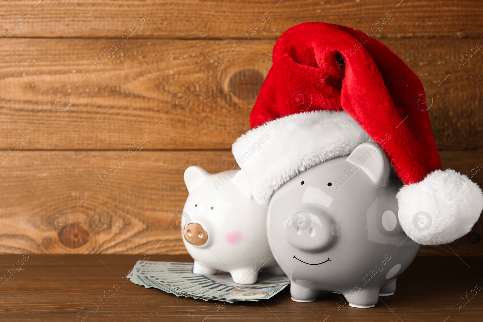 Photo of Piggy banks with Santa hat and dollar banknotes on wooden table. Space for text