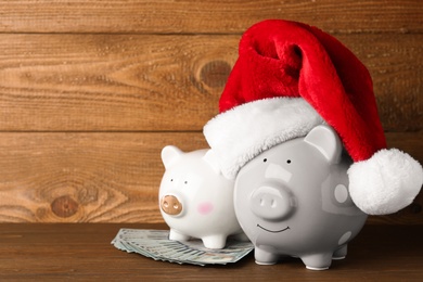 Photo of Piggy banks with Santa hat and dollar banknotes on wooden table. Space for text