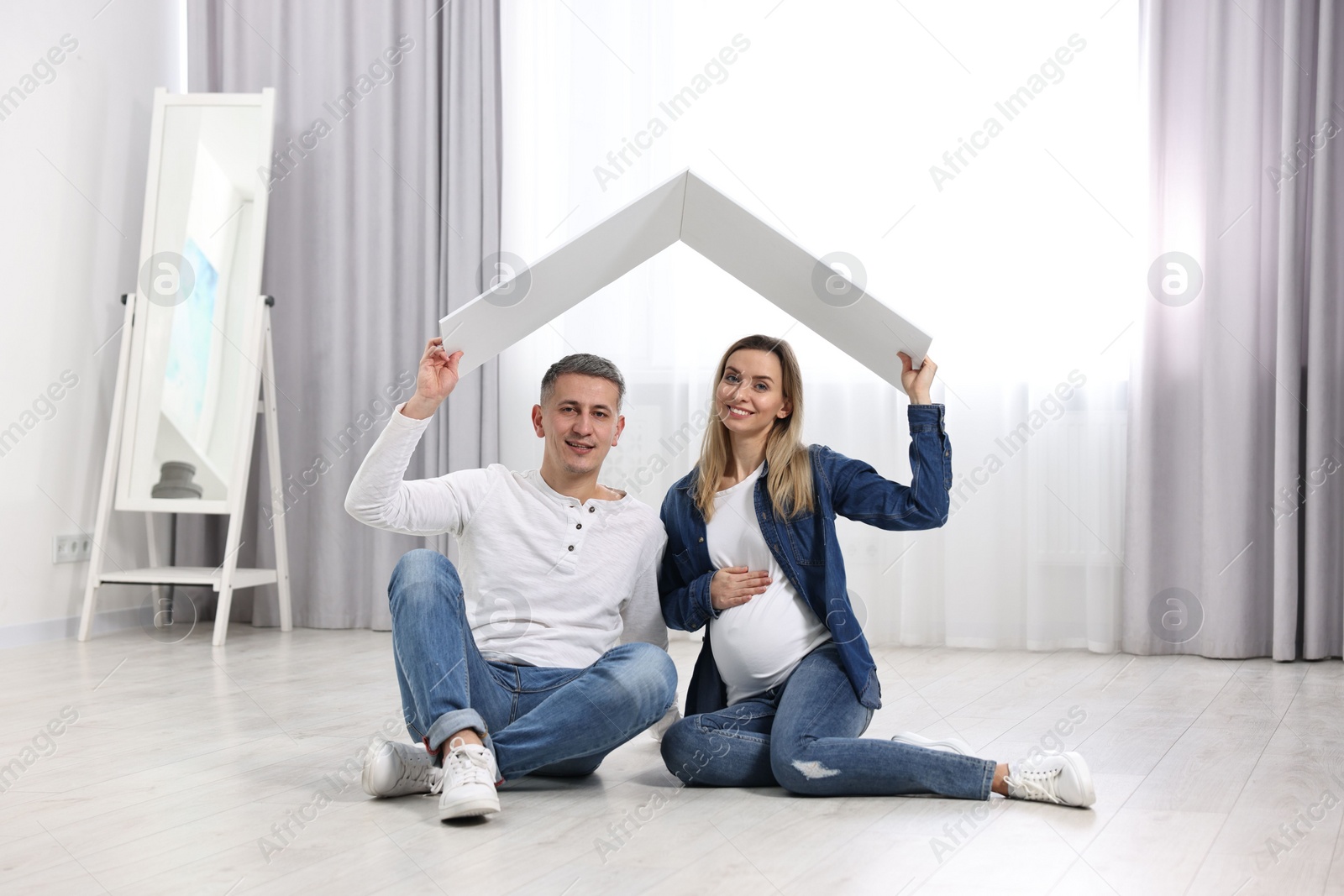 Photo of Young family housing concept. Pregnant woman with her husband sitting under cardboard roof on floor at home