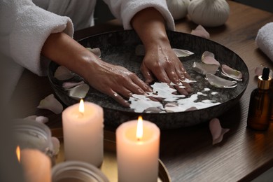 Photo of Woman soaking her hands in bowl of water and flower petals at wooden table, closeup. Spa treatment