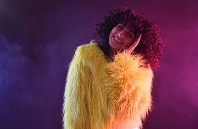 Beautiful young woman in yellow fur coat on color background in neon lights and smoke