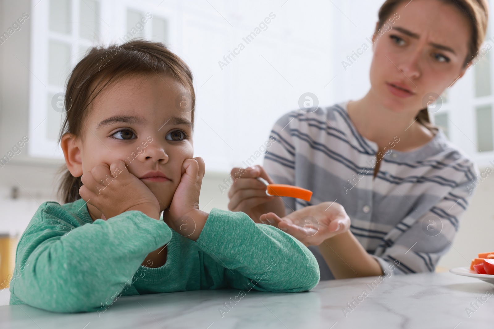 Photo of Mother feeding her daughter in kitchen. Little girl refusing to eat vegetables