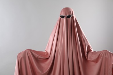Glamorous ghost. Woman in pink sheet with sunglasses on light grey background, space for text