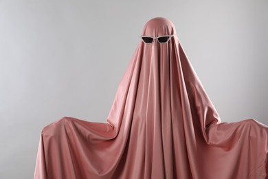 Photo of Glamorous ghost. Woman in pink sheet with sunglasses on light grey background, space for text