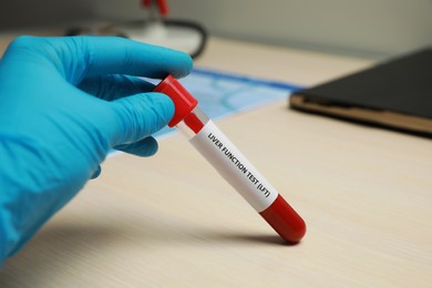 Laboratory worker holding tube with blood sample and label Liver Function Test at wooden table, closeup