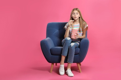 Emotional teenage girl with popcorn sitting in armchair during cinema show on color background