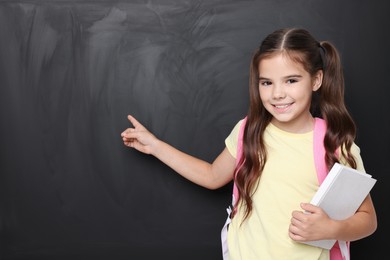 Photo of Cute schoolgirl with book near chalkboard, space for text