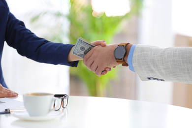 Photo of Man and woman shaking hands with dollars between their palms at table indoors, closeup. Bribery concept