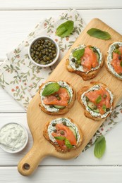 Tasty canapes with salmon, capers, cucumber and sauce on white wooden table, top view