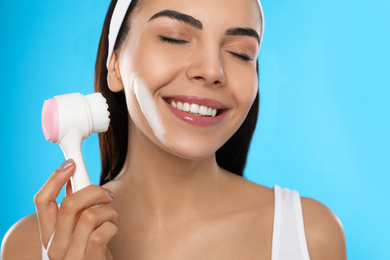 Photo of Young woman using facial cleansing brush on light blue background, closeup. Washing accessory