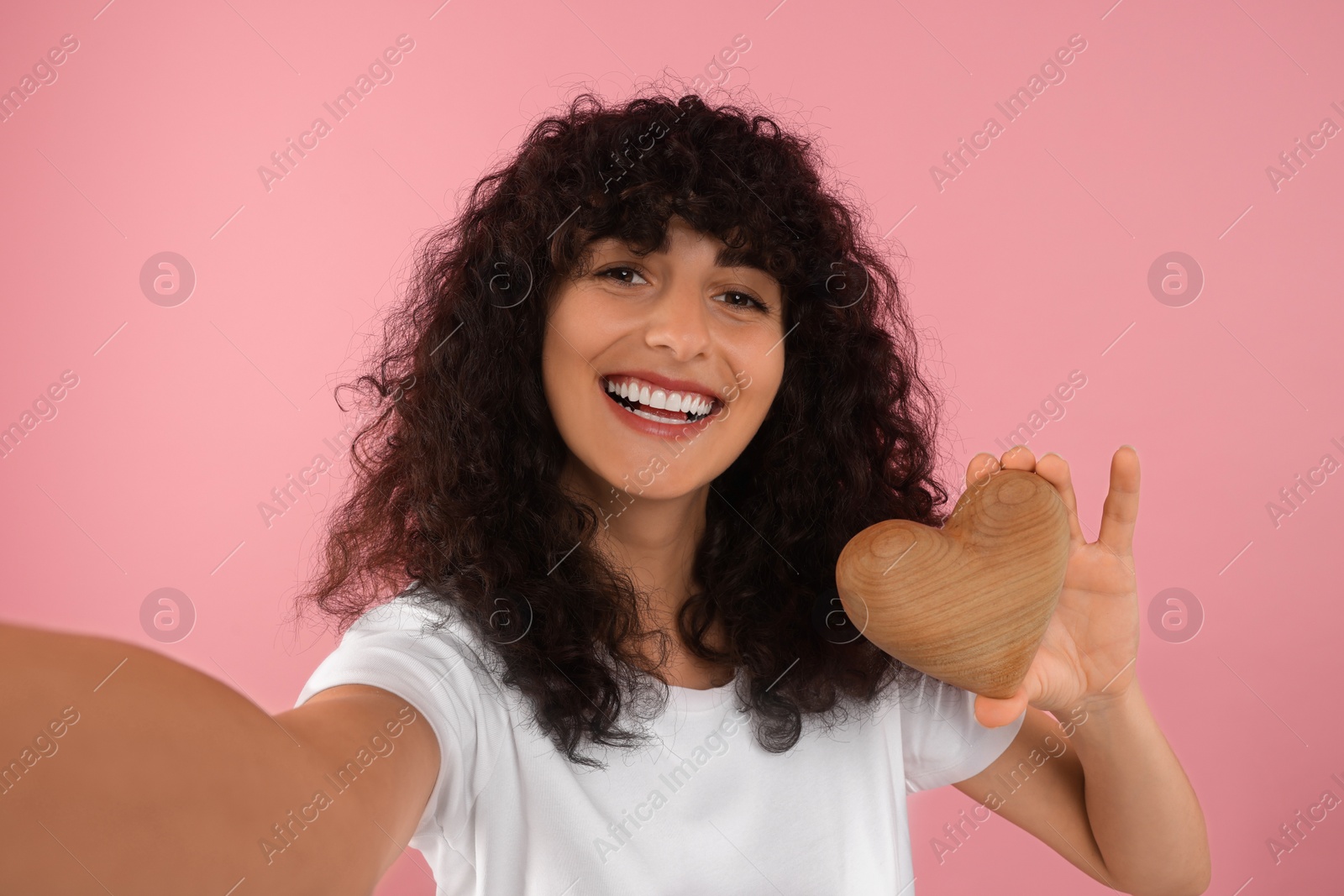 Photo of Happy young woman holding decorative wooden heart and taking selfie on pink background