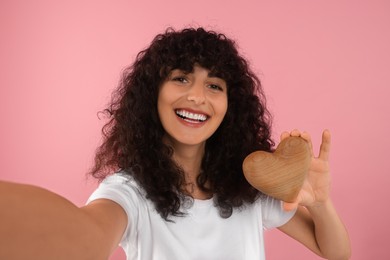 Happy young woman holding decorative wooden heart and taking selfie on pink background