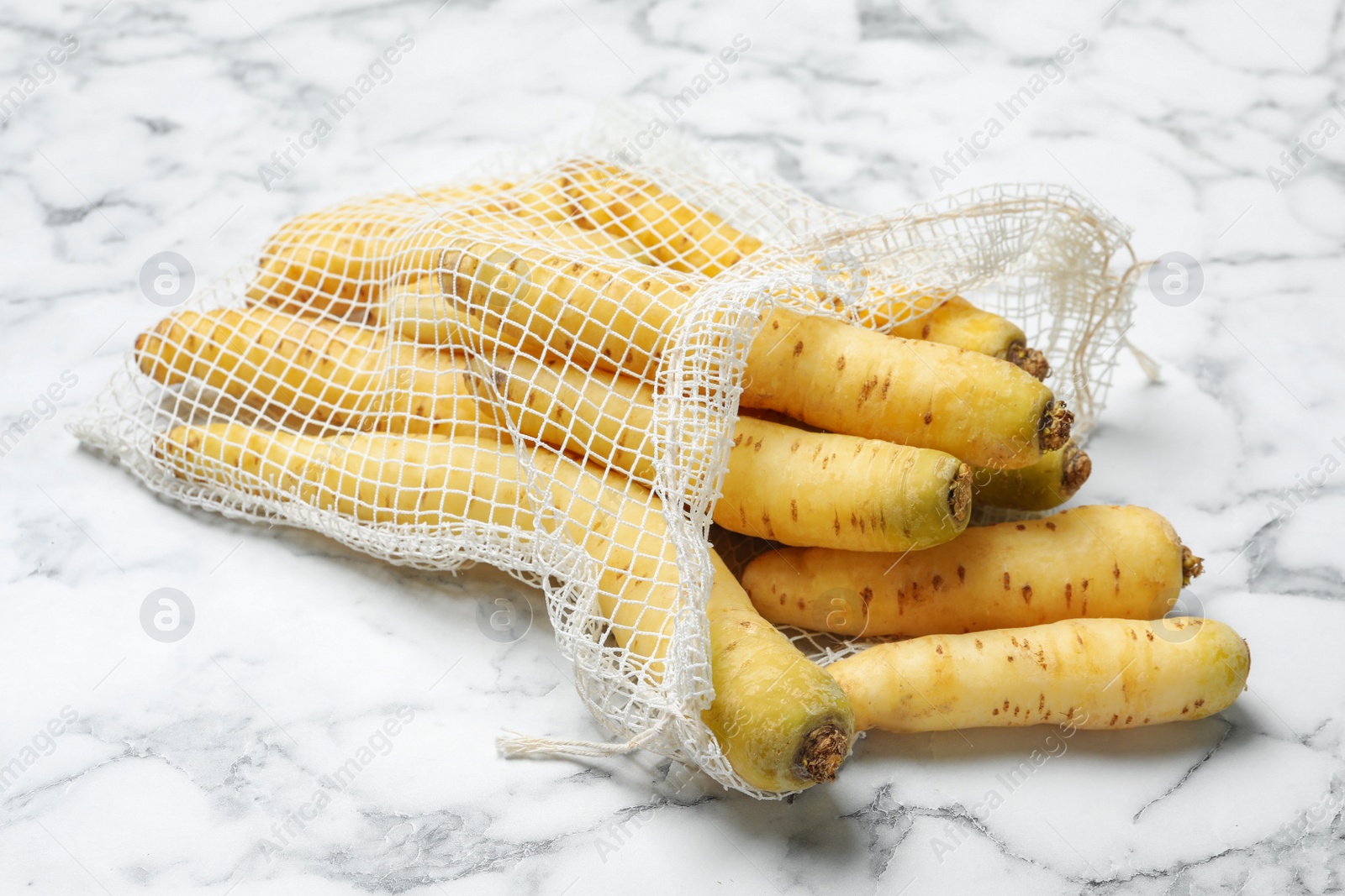 Photo of Many raw white carrots in mesh bag on marble table