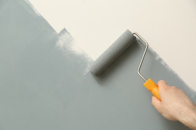 Photo of Man applying grey paint with roller brush on white wall, closeup