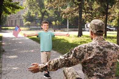 Photo of Little boy with flag of USA running towards his father in military uniform outdoors. Family reunion